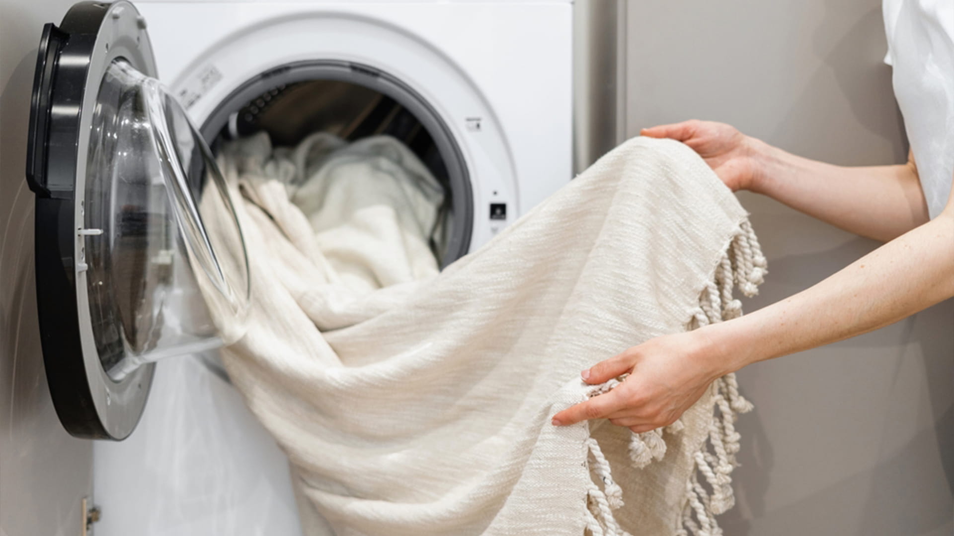  Close-up of hands gently removing a thick, woven blanket or throw with tassels from the open door of a washing machine, illustrating specialized care for household items.