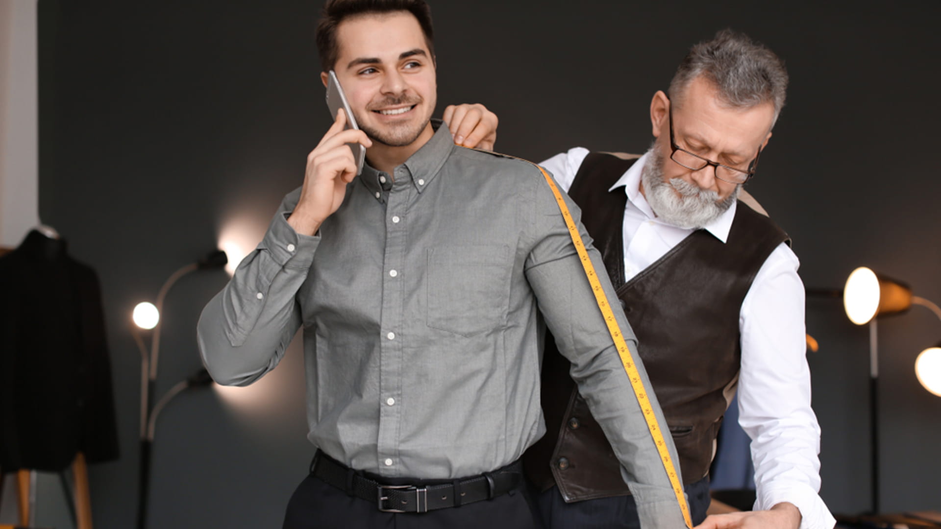 A senior tailor with a white beard and glasses uses a yellow tape measure to measure the sleeve of a smiling young man in a grey button-down shirt.
