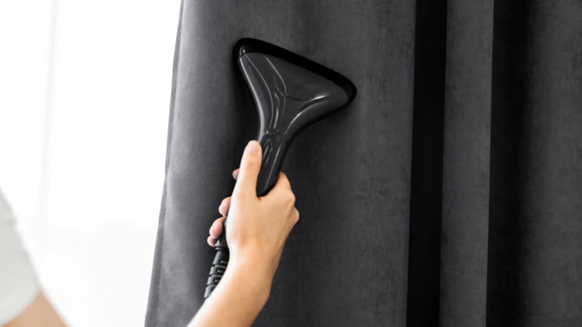 Handheld steamer used for steam cleaning a dark fabric curtain, removing wrinkles and refreshing the material.